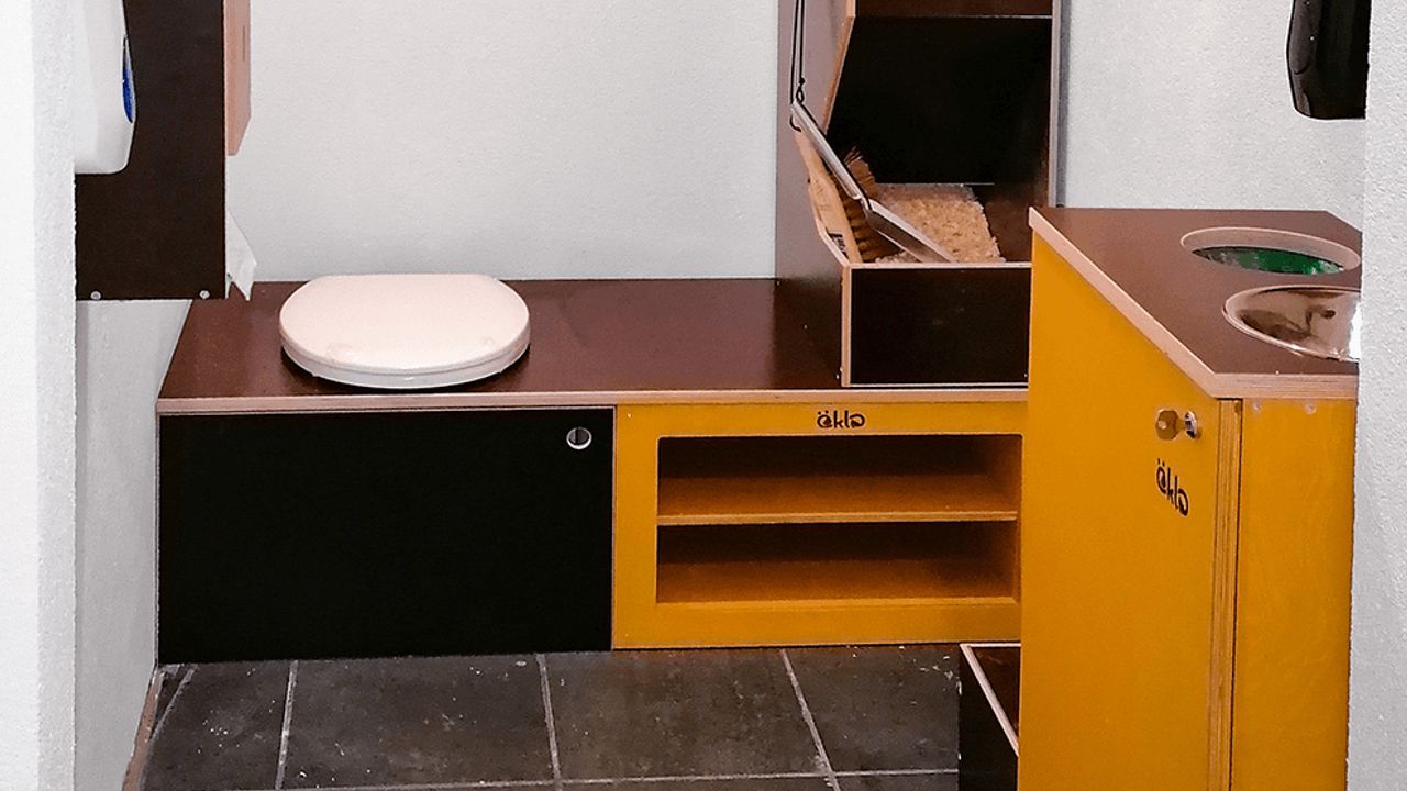 Ein öKlo, das in einem Kasten eingebaut in einem Toilettenraum steht. Im selben Raum ist eine öKlo Waschstation zu sehen. Trotz Anschlusspflicht können diese Produkte im Eigenheim installiert werden.