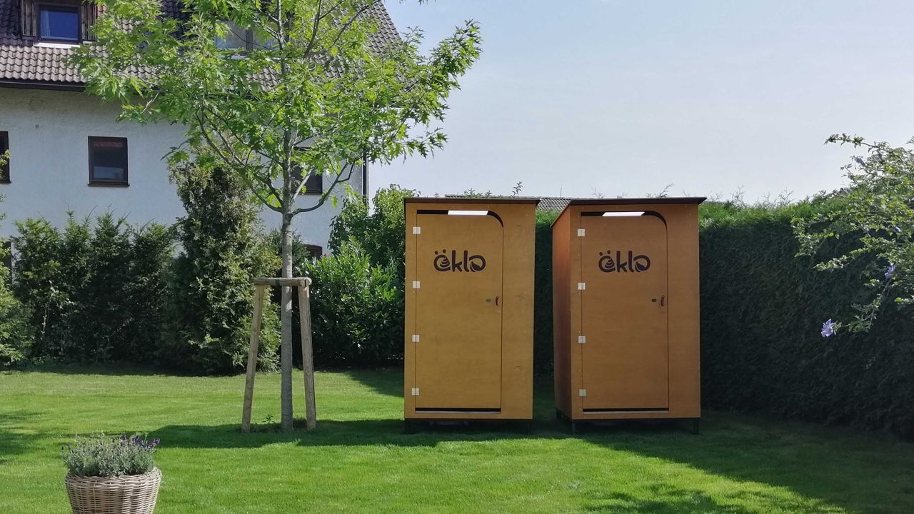 Zwei öKlo-Kabinen stehen nebeneinander auf einer grünen Wiese. Link neben ihnen steht ein junger Baum, rechts und hinter den Toiletten ist eine Buschwand zu sehen. Im linken Abschnitt des Bildes befindet sich hinter den Büschen ein weißes Haus mit braunem Wellziegeldach.