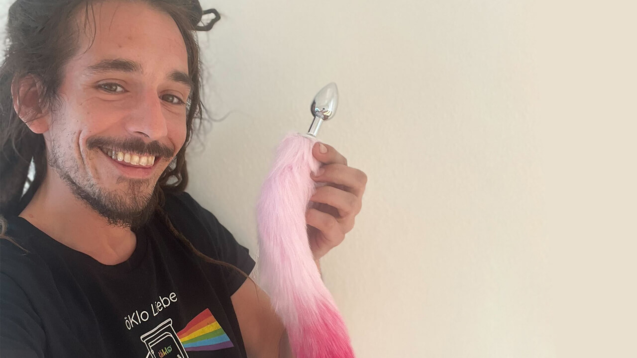 Lächelnder Mann mit öKlo-T-Shirt hält einen Analplug mit pinkfarbenen Plüschschwanz.