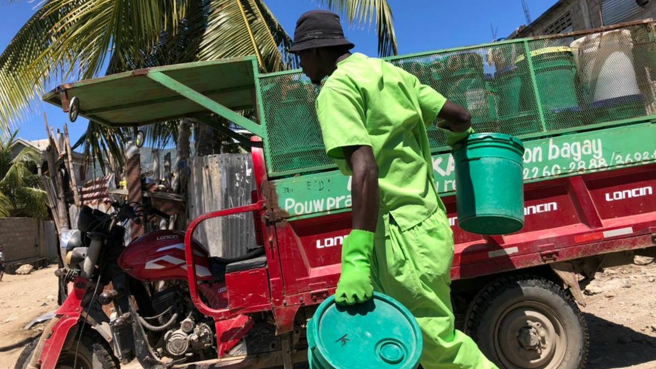 Eine Person mit dunkler Haut trägt ein Fass zu einem Fahrzeug. Sie trägt einen hellgrünen Anzug und einen dunklen Sonnenhut. Im Hintergrund sieht man Palmen.