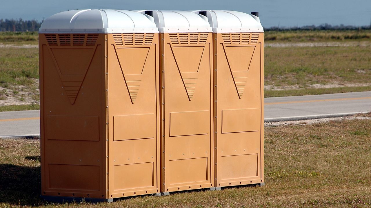 Drei Chemietoilettenkabinen aus Plastik stehen nebeneinander auf einer Wiese. Dahinter ist eine Straße zu sehen, hinter der noch mehr ebene Wiese liegt. Weit im Hintergrund ist ein Wald zu sehen. Die Kabinen sind orange-braun mit weißem, aufgewölbtem Dach.
